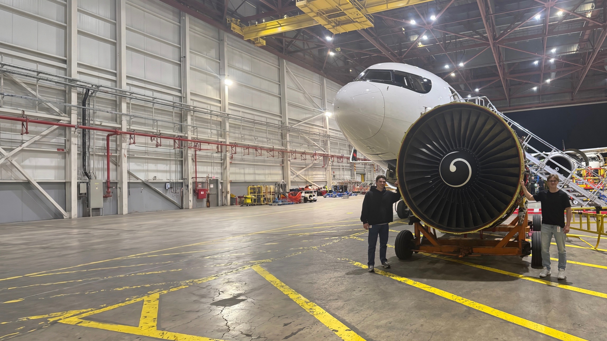 Two nuum robotics founders standing beside a CFM56 turbofan engine on a maintenance cart inside a wide aircraft hangar at night